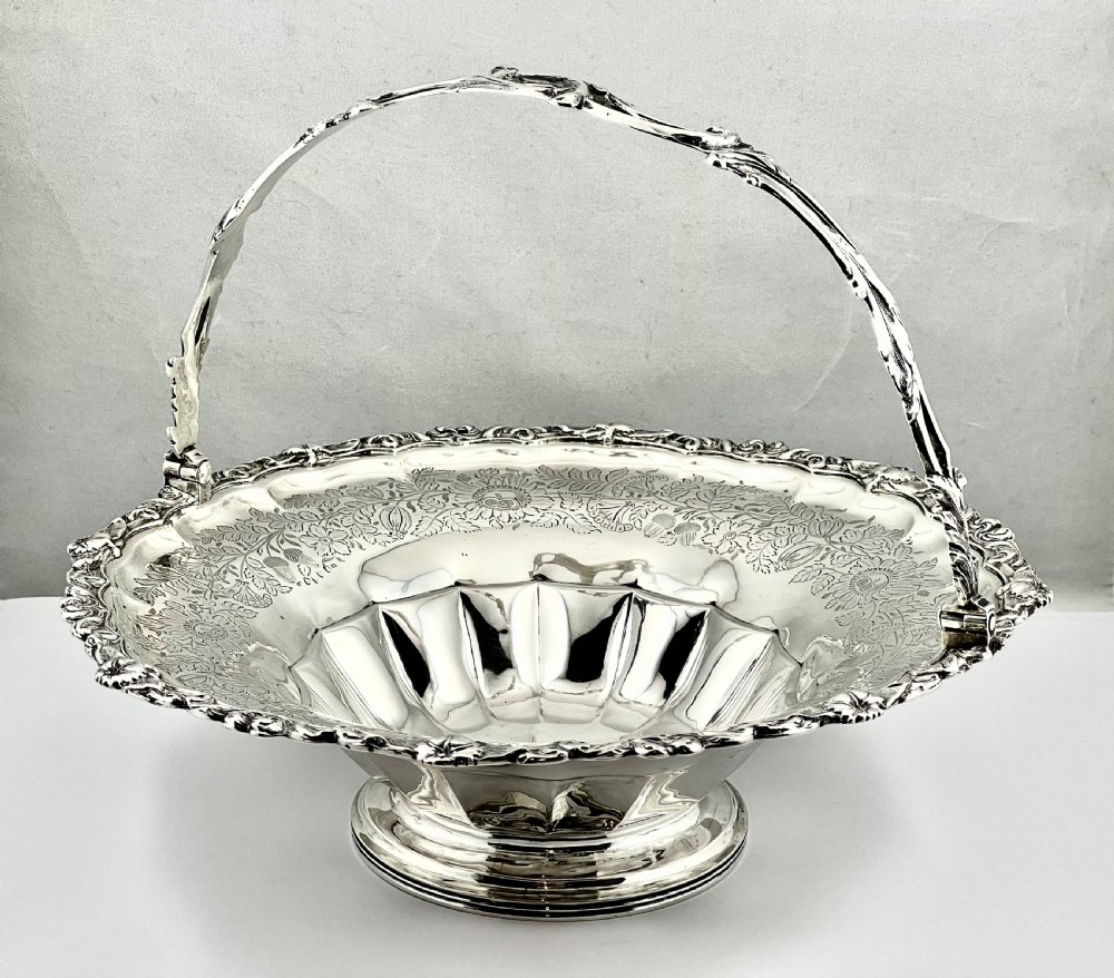 A round sterling silver bread basket featuring an overhead swing handle, floral engraving, a shell-detailed rim, and a fluted pedestal base.