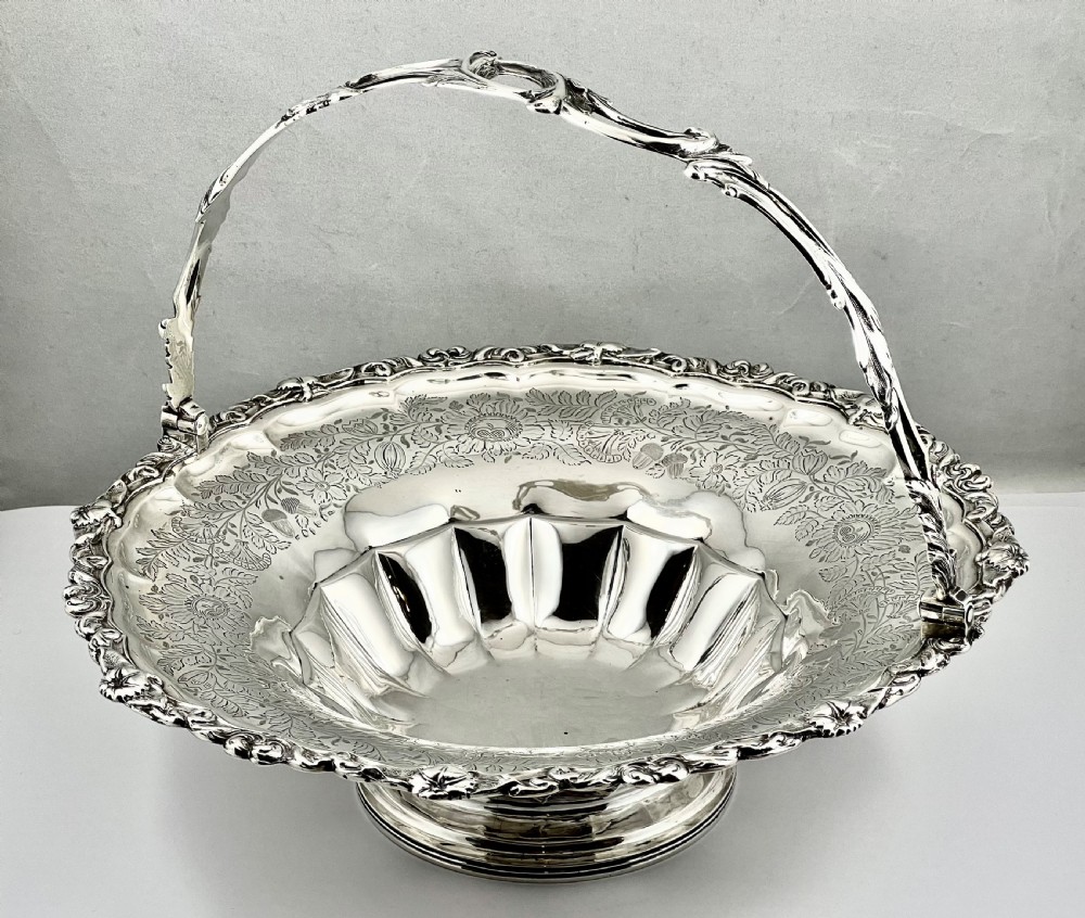 A round sterling silver bread basket featuring an overhead swing handle, floral engraving, a shell-detailed rim, and a fluted pedestal base. View II