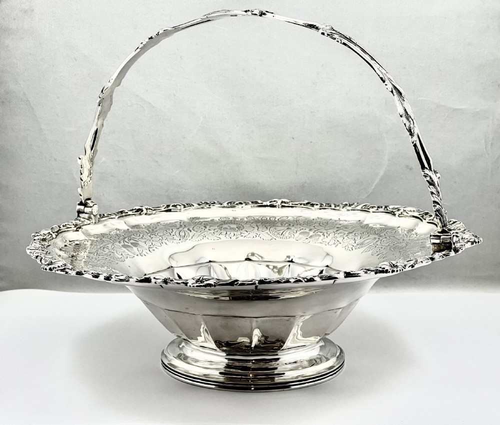 A round sterling silver bread basket featuring an overhead swing handle, floral engraving, a shell-detailed rim, and a fluted pedestal base. View III