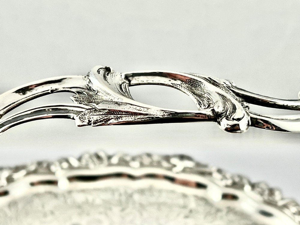 A round sterling silver bread basket featuring an overhead swing handle, floral engraving, a shell-detailed rim, and a fluted pedestal base. View VII