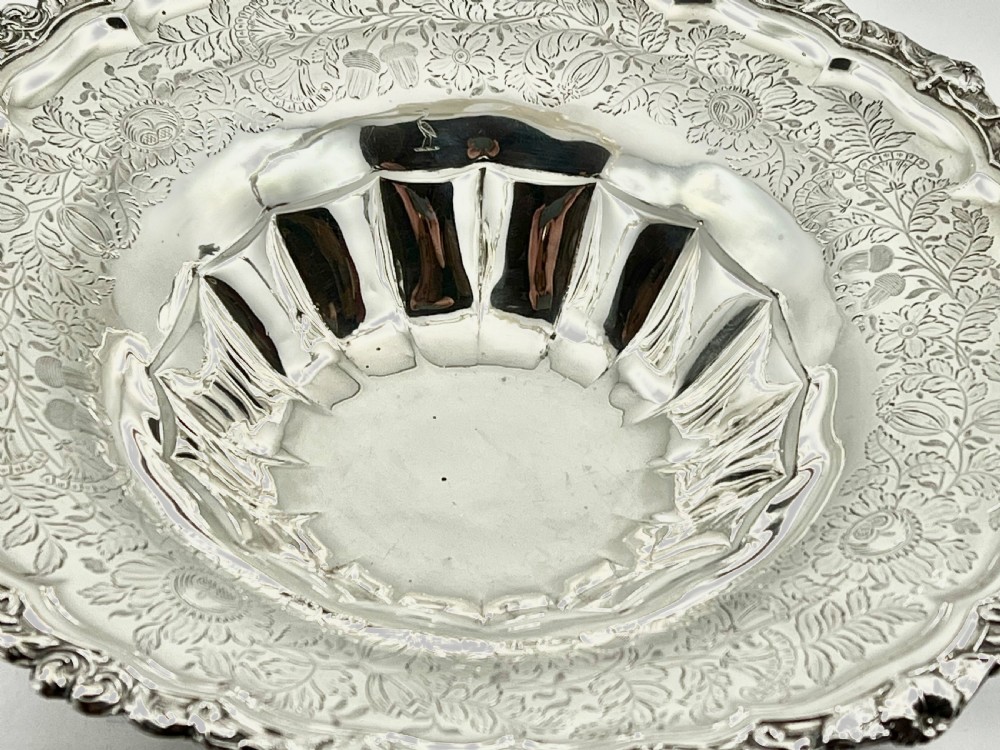 A round sterling silver bread basket featuring an overhead swing handle, floral engraving, a shell-detailed rim, and a fluted pedestal base. View X