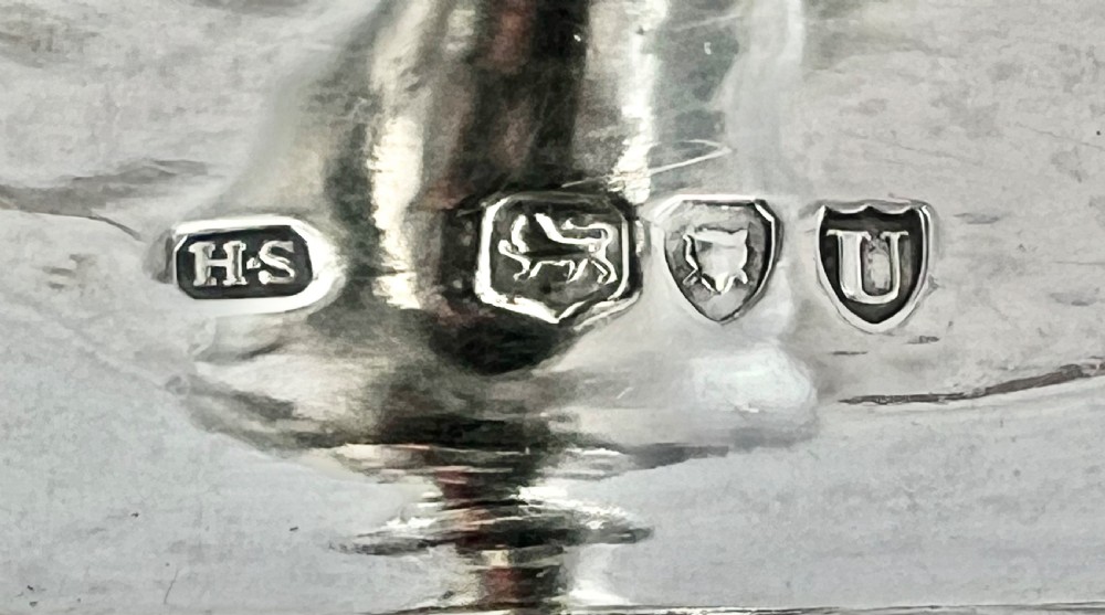 A Victorian sterling silver chamber stick with a square tray base, gadrooned borders, a scroll handle with an engraved crest, and a conical snuffer. View X