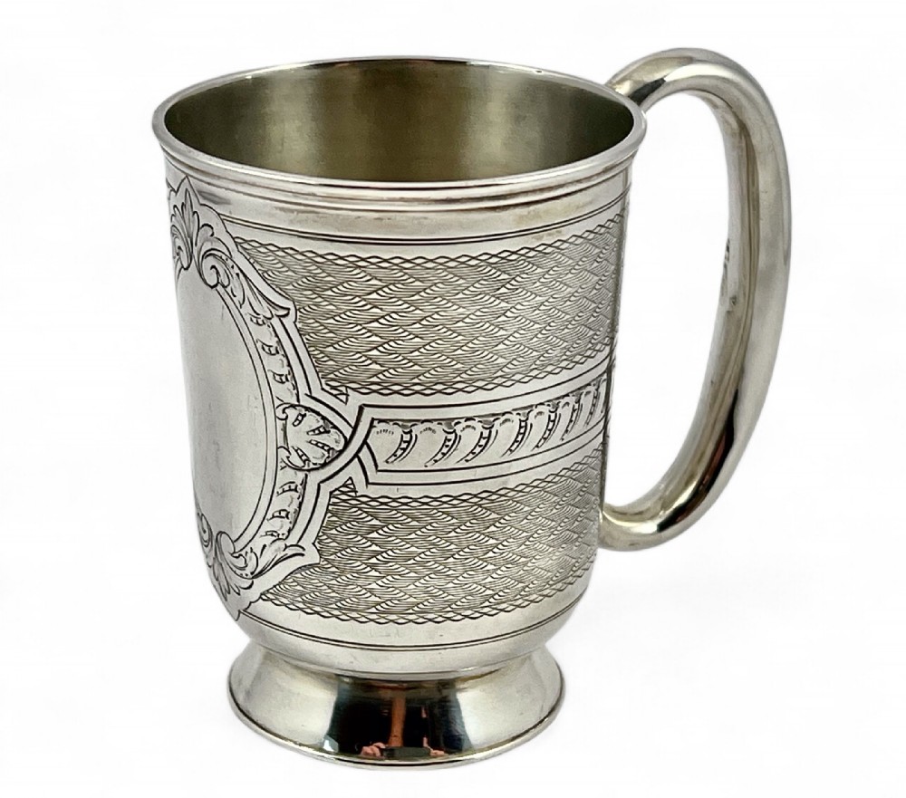 A Victorian sterling silver christening cup featuring detailed engine-turned geometric engraving, an oval cartouche, and a loop handle resting on a circular foot.