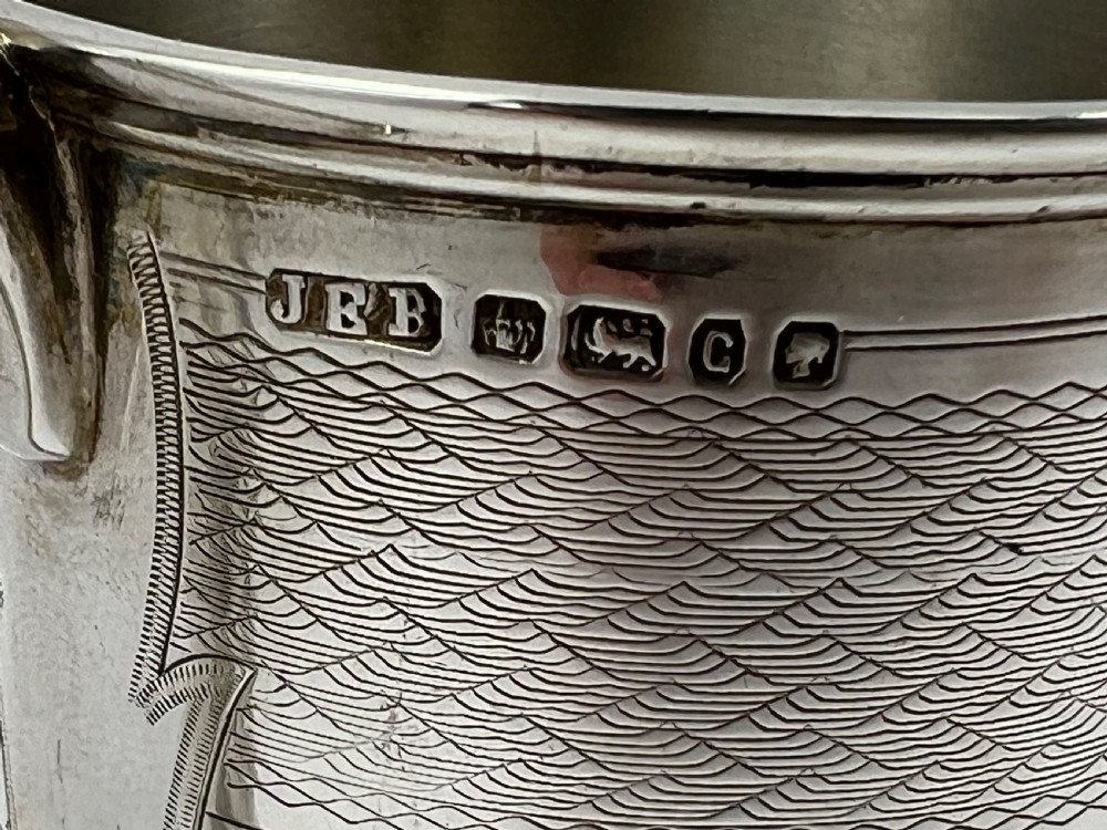 A Victorian sterling silver christening cup featuring detailed engine-turned geometric engraving, an oval cartouche, and a loop handle resting on a circular foot. View VIII