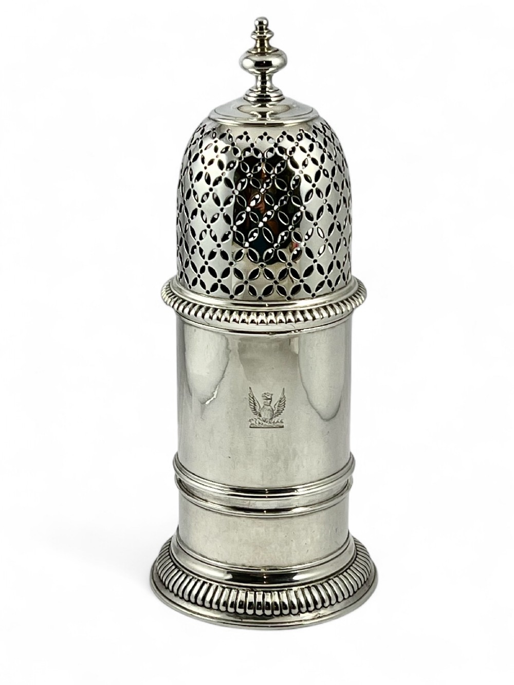 A cylindrical antique sterling silver sugar caster featuring a pierced dome lid, finial top, reeded borders, and an engraved armorial crest on the body.