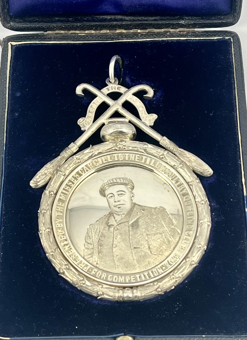 An Edwardian silver oval curling medal featuring crossed brooms at the top, a central engraved portrait of a man, and an inscribed silver border. View IX