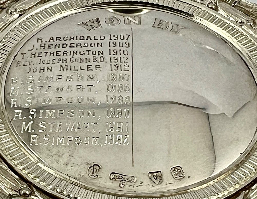 An Edwardian silver oval curling medal featuring crossed brooms at the top, a central engraved portrait of a man, and an inscribed silver border. View XII