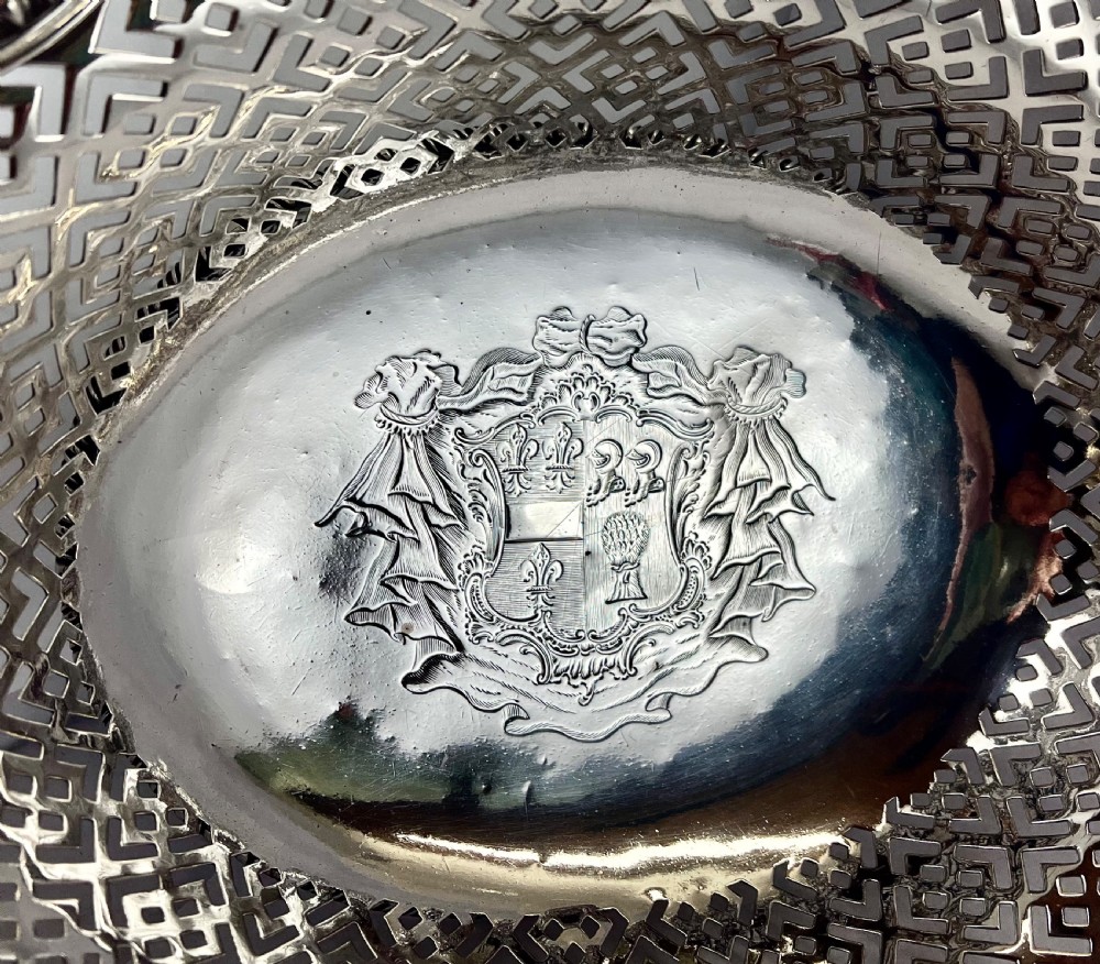 An 18th-century George II oval sterling silver basket featuring hand-pierced fretwork sides, an engraved central coat of arms, and an overhead swing handle. View VII