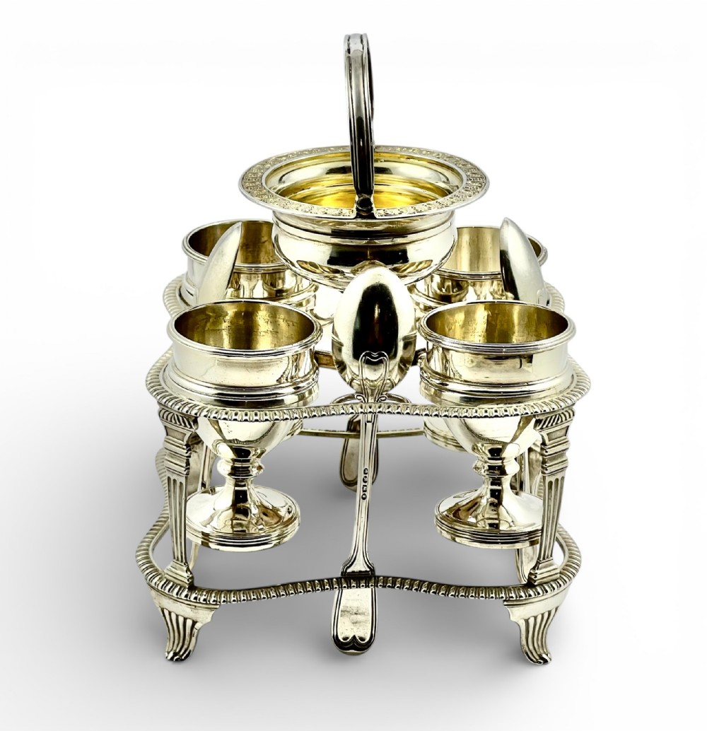 A George III sterling silver egg cruet set featuring a central handle, a gilt-lined salt cellar, six egg cups, and fitted silver spoons on a gadrooned stand.