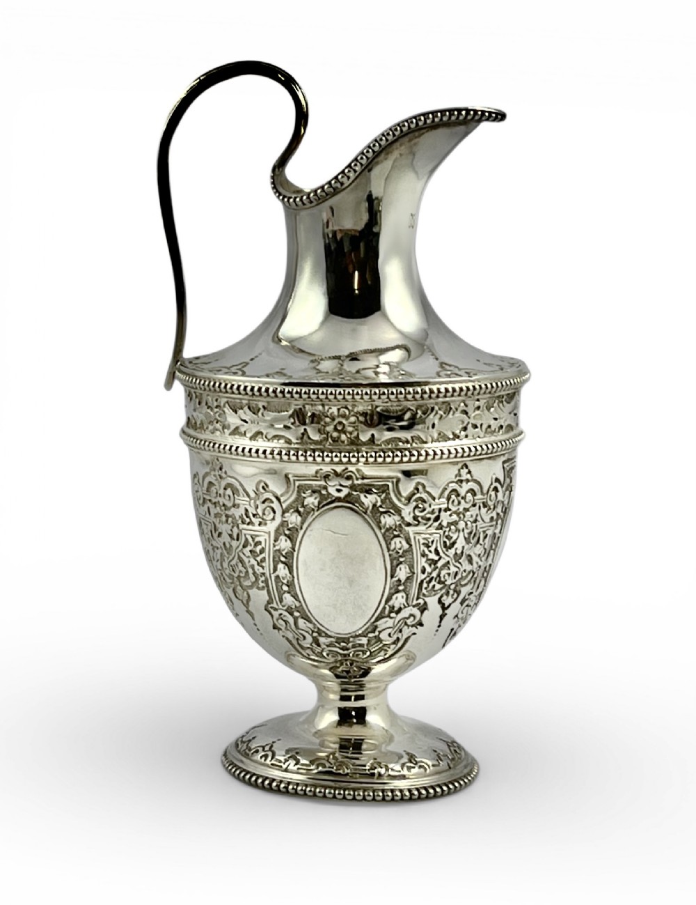 A Victorian sterling silver pedestal cream jug featuring intricate engraved scrollwork, a blank oval cartouche, a beaded rim, and a high loop handle.