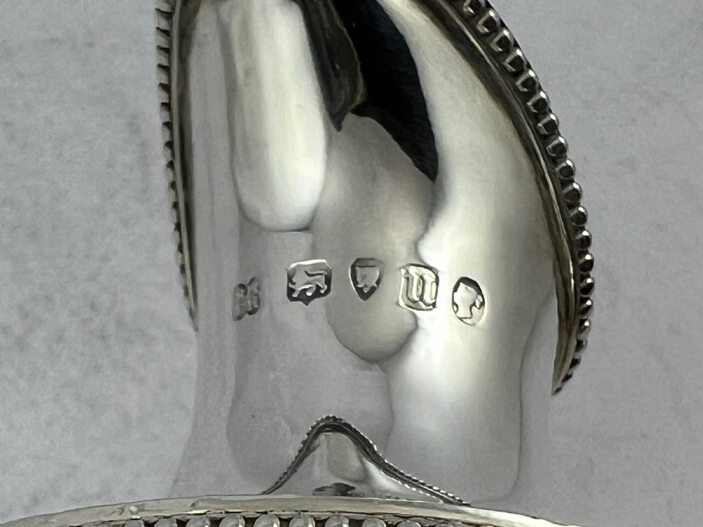 A Victorian sterling silver pedestal cream jug featuring intricate engraved scrollwork, a blank oval cartouche, a beaded rim, and a high loop handle. View VII