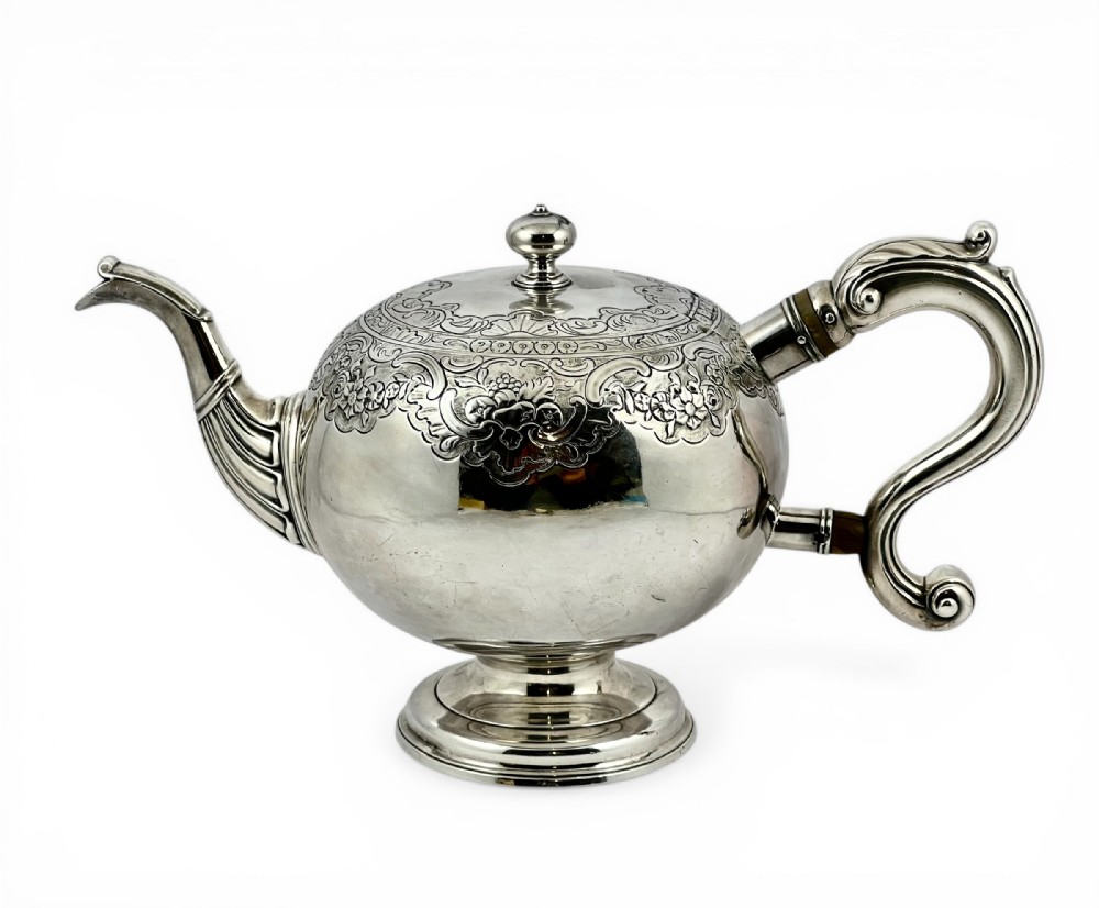 A George II Scottish sterling silver bullet teapot featuring chased foliage along the shoulder, a wooden scroll handle, and a raised pedestal base.