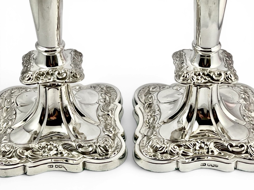 A pair of polished sterling silver candlesticks featuring tapered columns, ornate scroll-patterned square bases, and hallmarked edges. View V