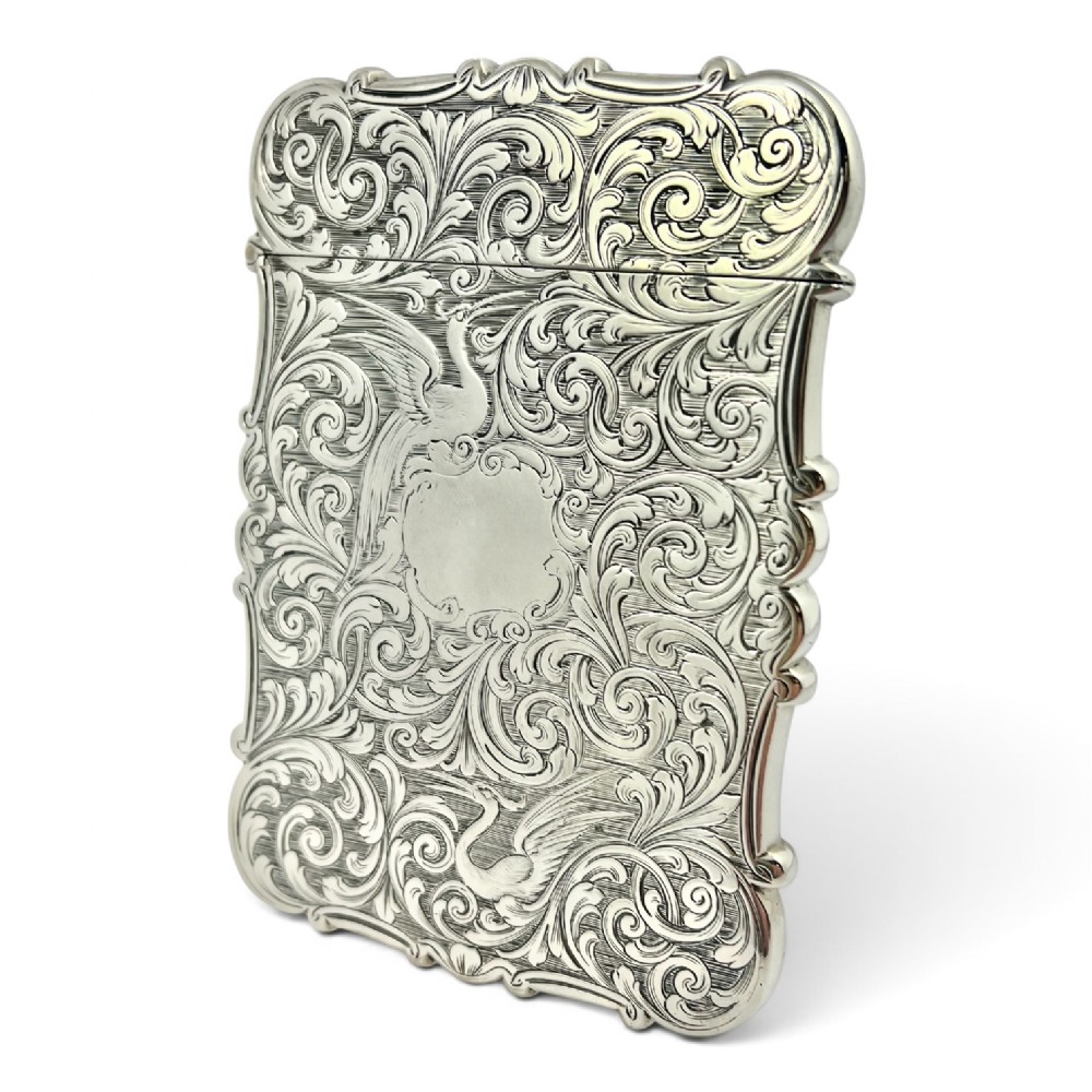 A Victorian sterling silver card case with detailed foliate scrollwork engraving, bird motifs, a blank central cartouche, and a contoured edge.