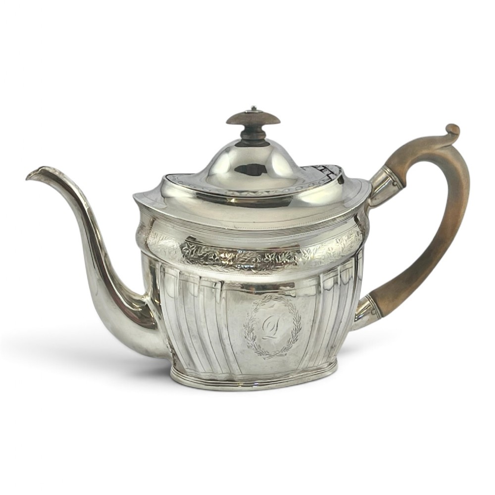 An oval Georgian sterling silver teapot featuring fluted lower body panelling, engraved decorative borders, a curved wooden handle, and a matching silver lid with a wood finial.