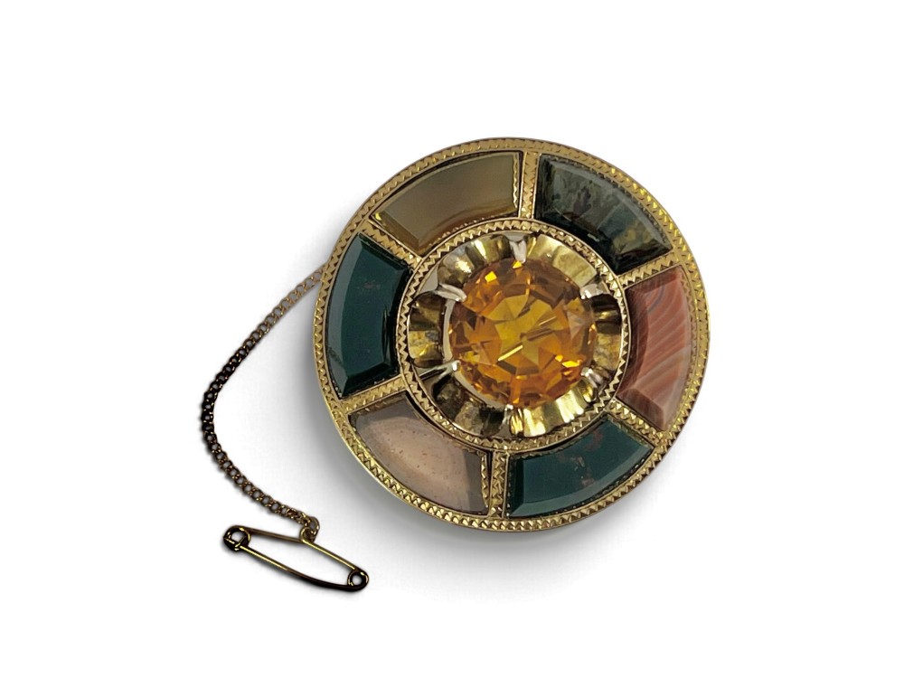 A circular Victorian 18ct gold brooch set with a central faceted yellow citrine gemstone surrounded by wedge-shaped inlaid agate and bloodstone panels, fitted with a safety chain. View VI