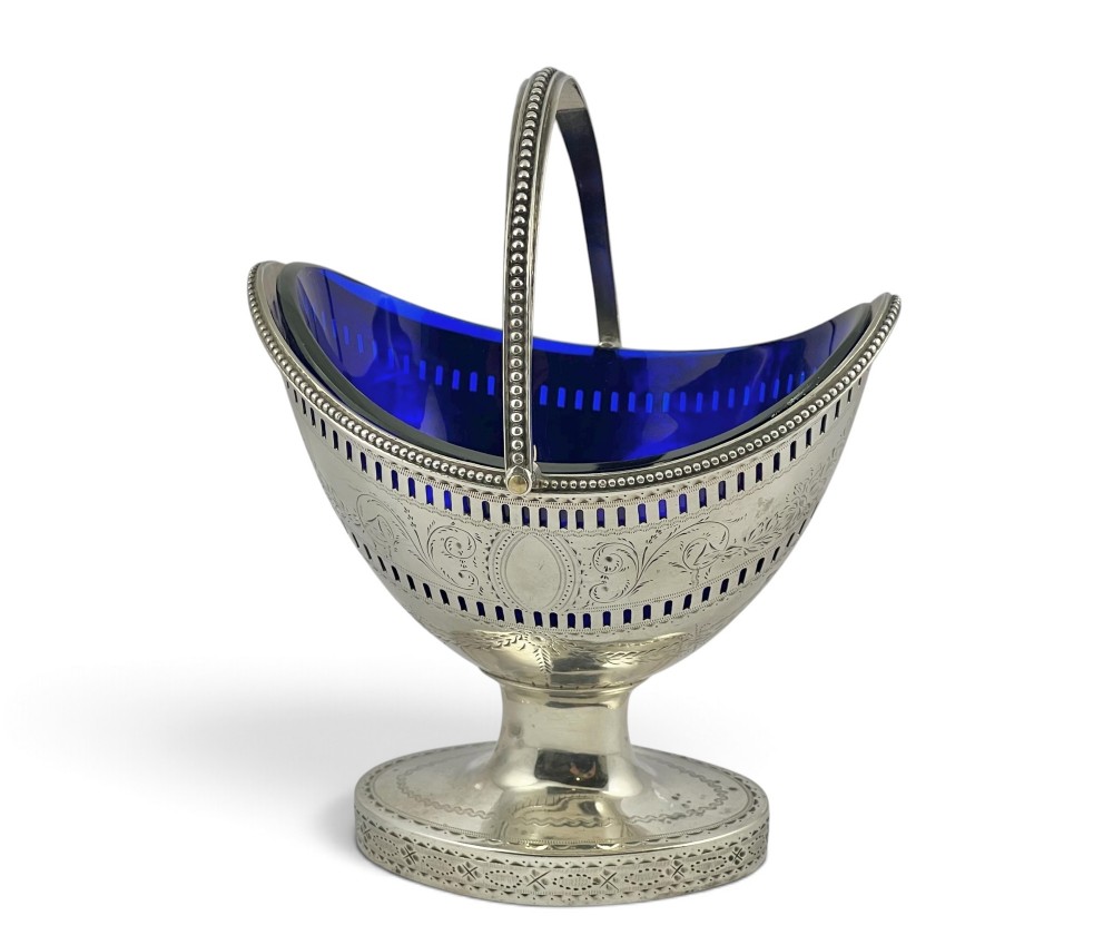 A George III sterling silver sugar basket featuring pierced geometric engraving, a beaded swing handle, a pedestal base, and a fitted cobalt blue glass liner.
