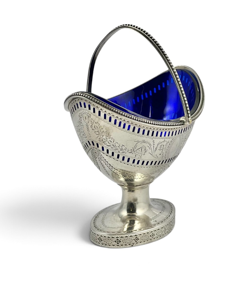 A George III sterling silver sugar basket featuring pierced geometric engraving, a beaded swing handle, a pedestal base, and a fitted cobalt blue glass liner. View II