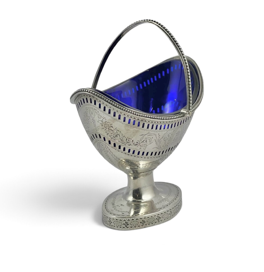 A George III sterling silver sugar basket featuring pierced geometric engraving, a beaded swing handle, a pedestal base, and a fitted cobalt blue glass liner. View IV