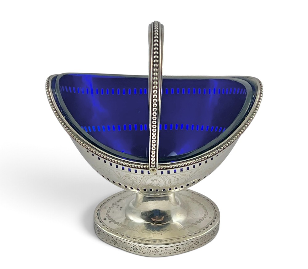 A George III sterling silver sugar basket featuring pierced geometric engraving, a beaded swing handle, a pedestal base, and a fitted cobalt blue glass liner. View V