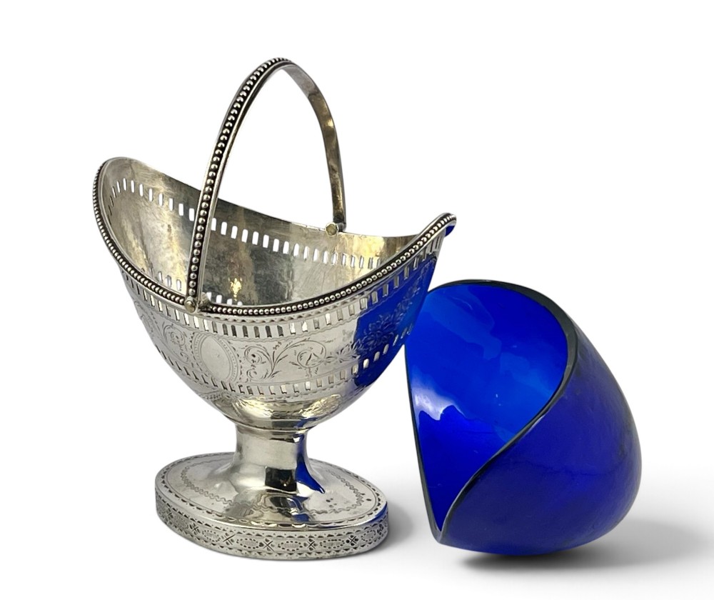 A George III sterling silver sugar basket featuring pierced geometric engraving, a beaded swing handle, a pedestal base, and a fitted cobalt blue glass liner. View VIII