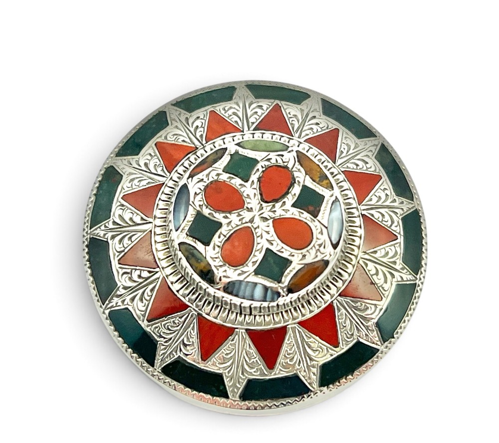 A circular Victorian Scottish silver brooch inlaid with geometric red carnelian, green jasper, and agate stones surrounding a central quatrefoil motif.