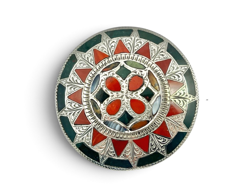 A circular Victorian Scottish silver brooch inlaid with geometric red carnelian, green jasper, and agate stones surrounding a central quatrefoil motif. View V