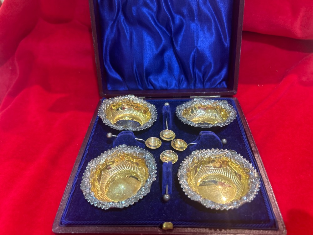 A set of four die-stamped silver open salt cellars with gilt interiors and matching salt spoons, arranged inside a fitted velvet-lined presentation case.
