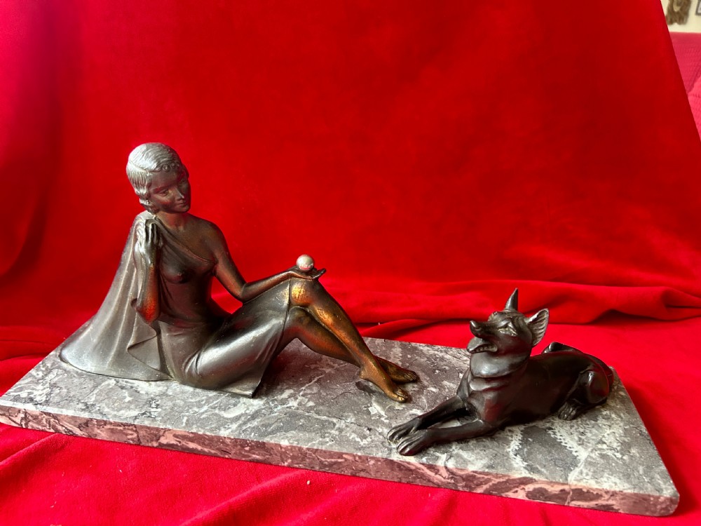 An Art Deco bronze figure group depicting a reclining stylized woman holding an orb beside a seated dog, set on a grey marble plinth.
