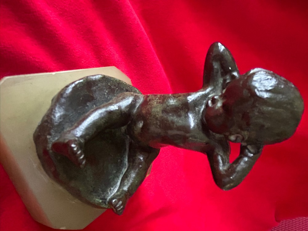 A patinated bronze figurine depicting a seated baby stretching with hands behind its head, mounted on a chamfered pale onyx base. View VII