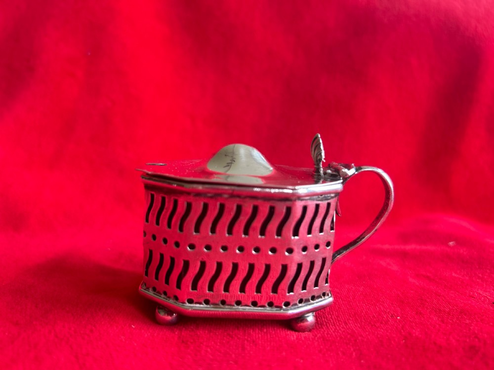 A pierced silver mustard pot featuring a hinged lid with thumbpiece, scroll handle, and small ball feet.
