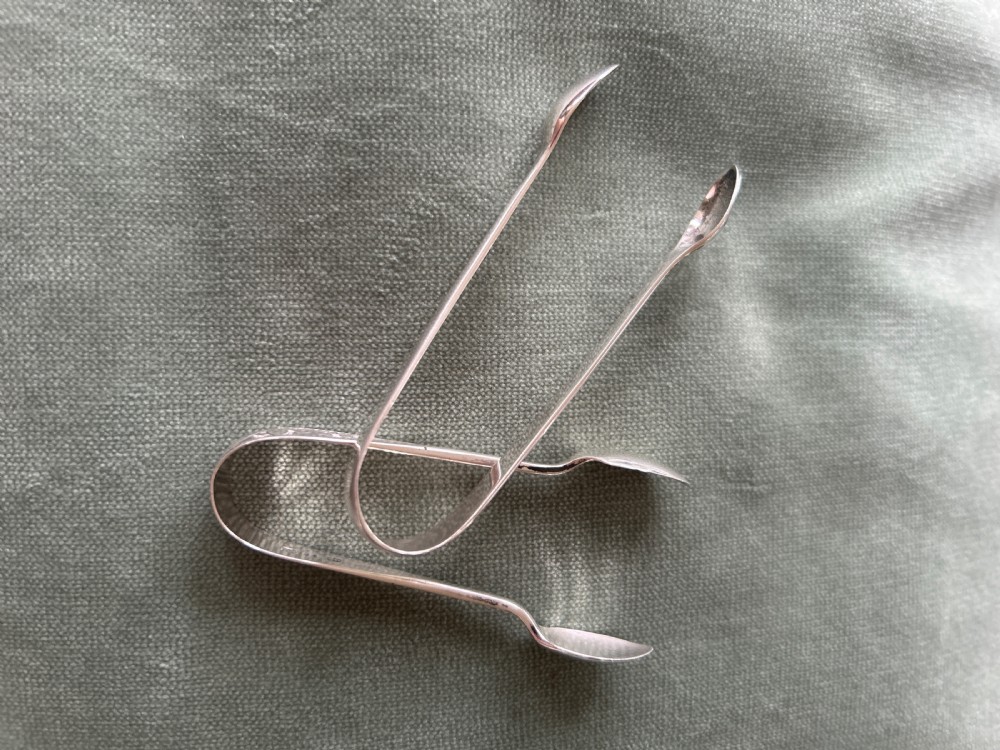 A pair of polished silver sugar tongs with spoon-shaped grips placed on a green fabric surface.
