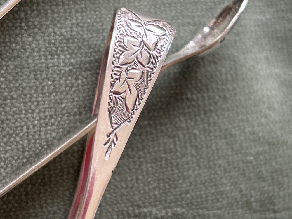 A pair of polished silver sugar tongs with spoon-shaped grips placed on a green fabric surface. View II