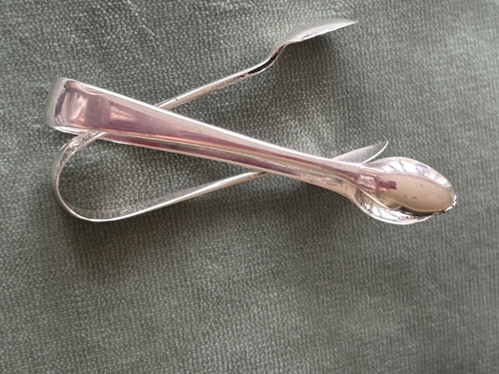 A pair of polished silver sugar tongs with spoon-shaped grips placed on a green fabric surface. View III