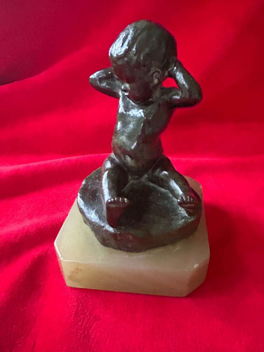 A patinated bronze figurine depicting a seated baby stretching with hands behind its head, mounted on a chamfered pale onyx base.