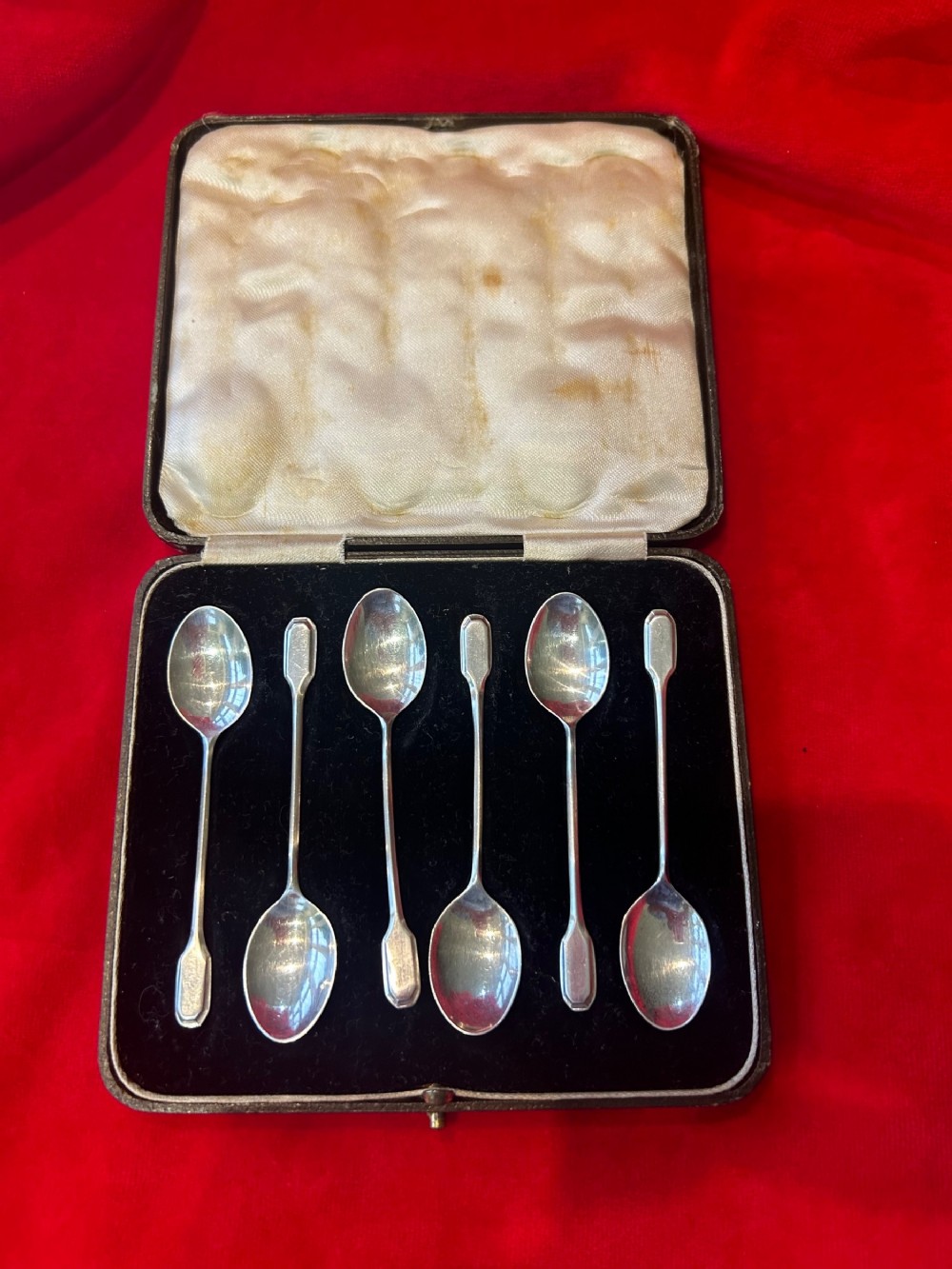 A set of six silver coffee spoons with simple geometric stems resting inside a black velvet and white satin lined presentation case.