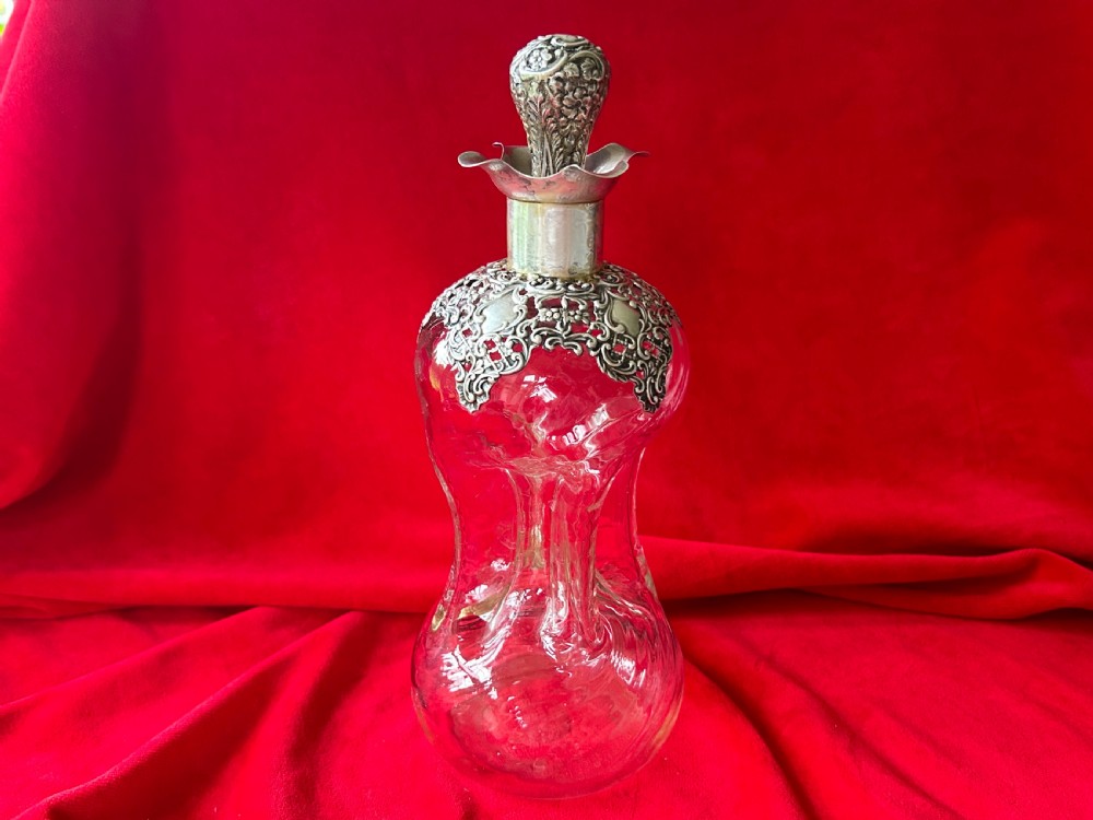 An Edwardian clear glass decanter featuring a pinched waist, decorative pierced silver overlay around the shoulder, and an ornate silver stopper.