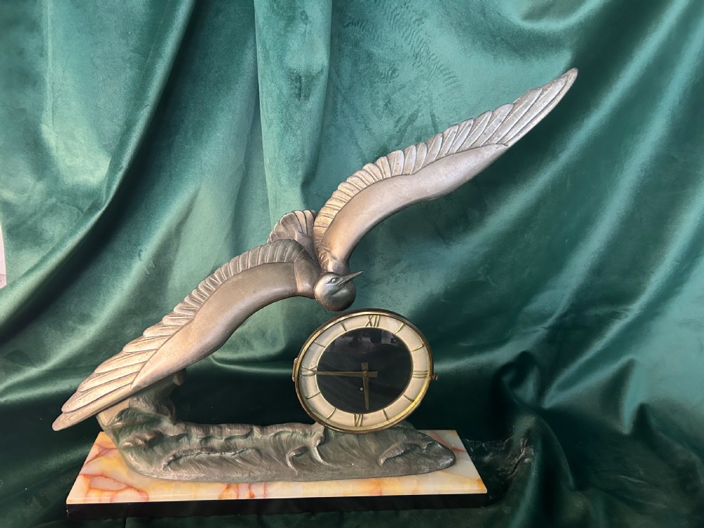 An Art Deco mantel clock featuring a metal bird figure in flight over waves beside a round dial on an onyx plinth.