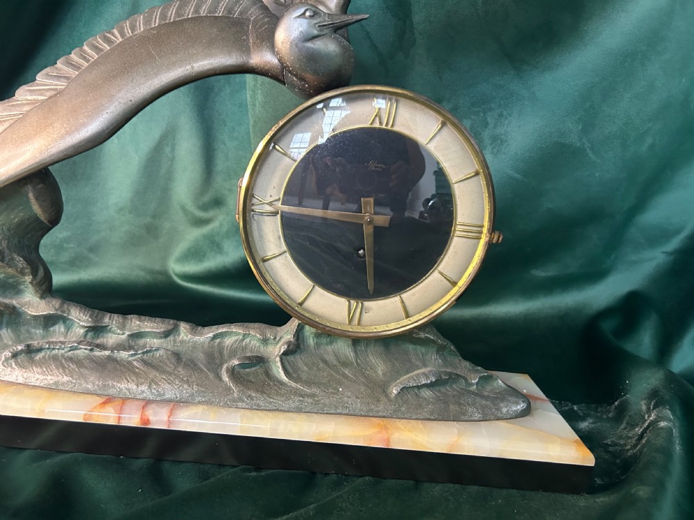 An Art Deco mantel clock featuring a metal bird figure in flight over waves beside a round dial on an onyx plinth. View II