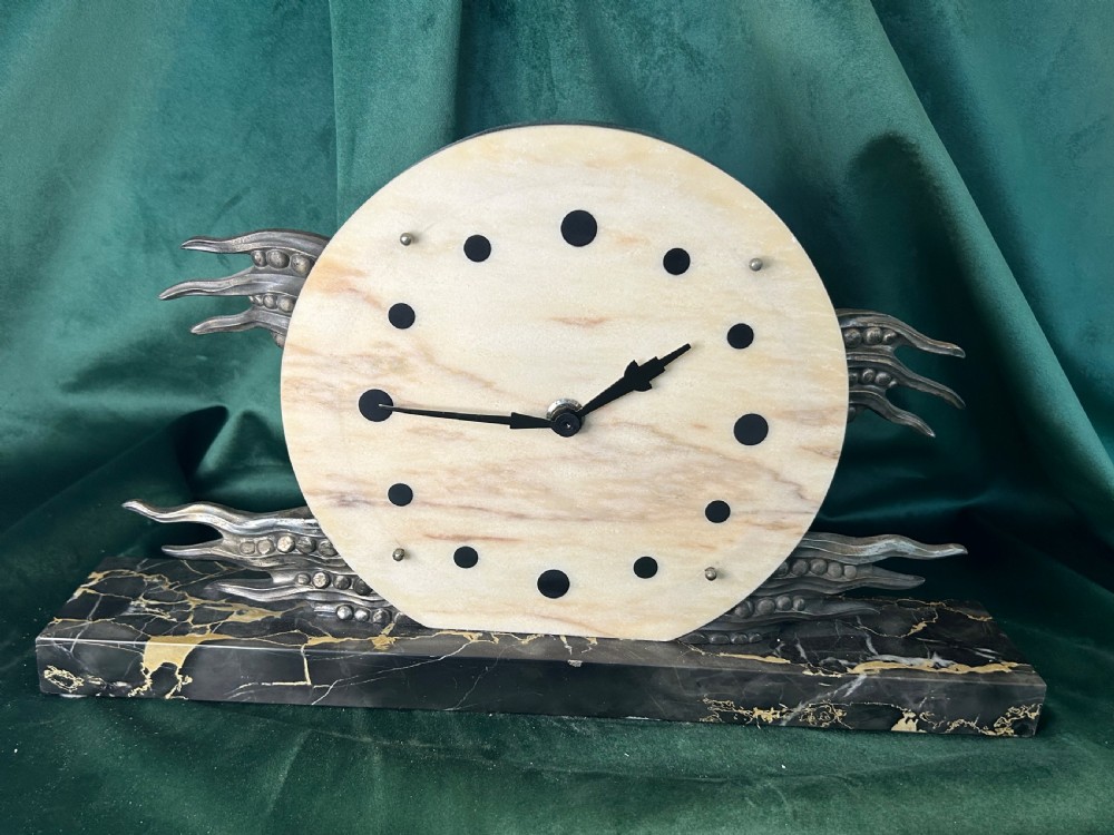 An Art Deco mantel clock featuring a circular cream alabaster dial with black circular hour markers, set upon a black veined marble base with silver-toned metal winged accents.