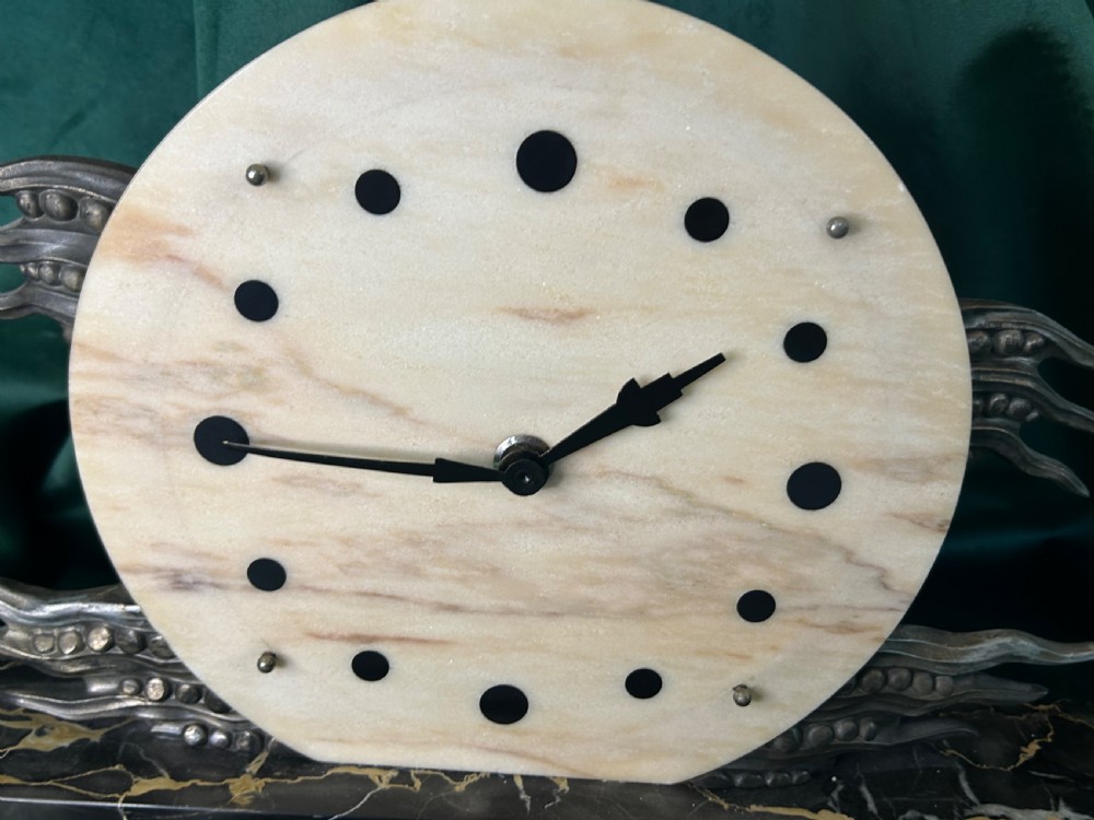 An Art Deco mantel clock featuring a circular cream alabaster dial with black circular hour markers, set upon a black veined marble base with silver-toned metal winged accents. View II