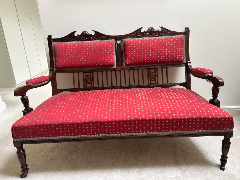 A Victorian mahogany settee featuring carved crest rails, twin padded back cushions in red patterned fabric, turned legs on castors, and a wide upholstered seat.