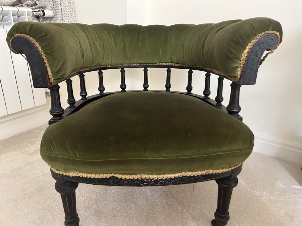 A Victorian ebonised wood tub armchair featuring olive green velvet upholstery, a padded horseshoe backrest supported by turned spindles, and gold braided trim.
