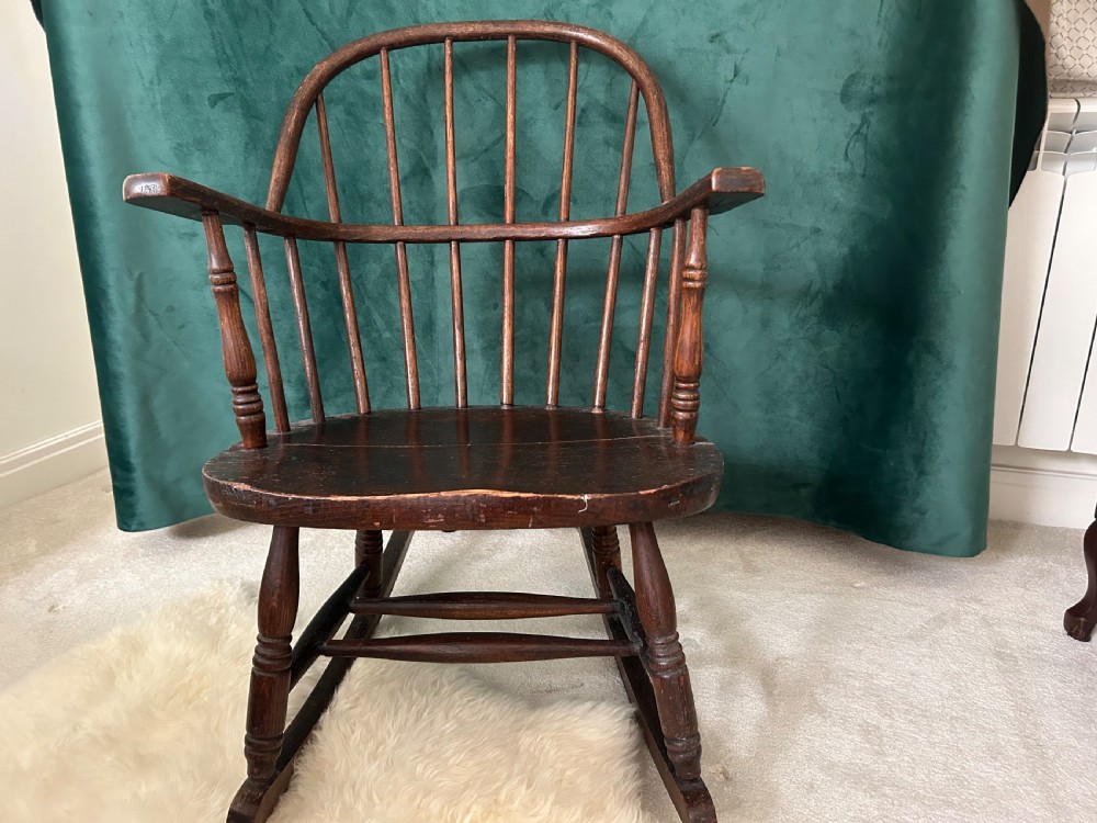 A dark stained wooden Windsor rocking chair featuring a spindle hoop back, curved armrests, turned legs, and wooden runners.