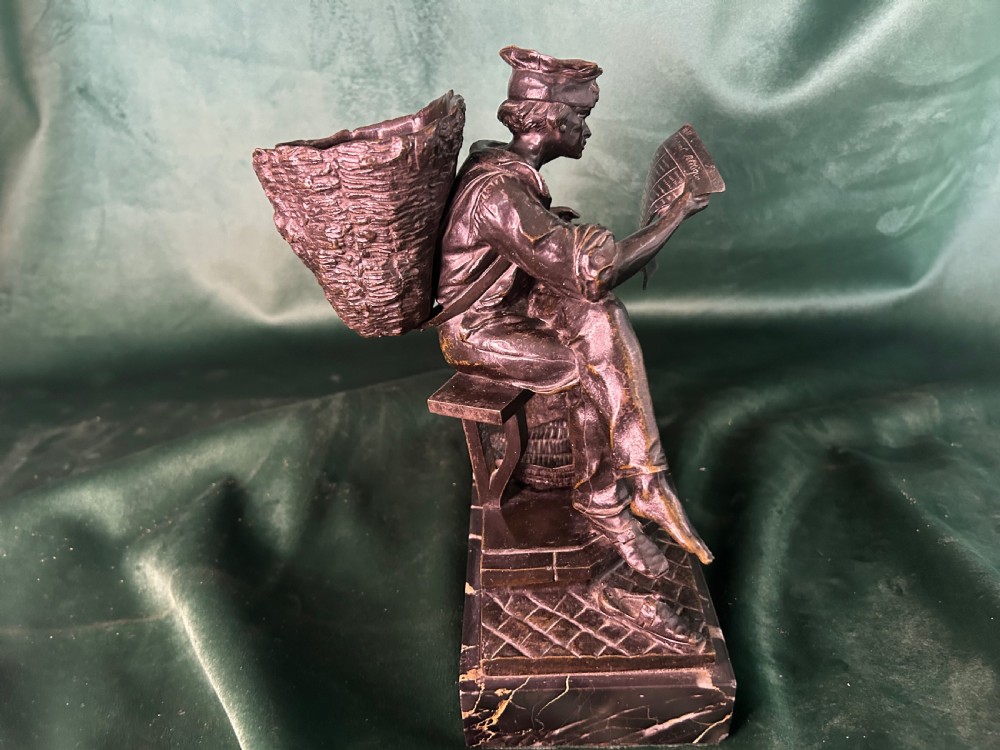 A patinated dark bronze sculpture depicting a seated man reading a newspaper, mounted on a black veined marble base. View IV