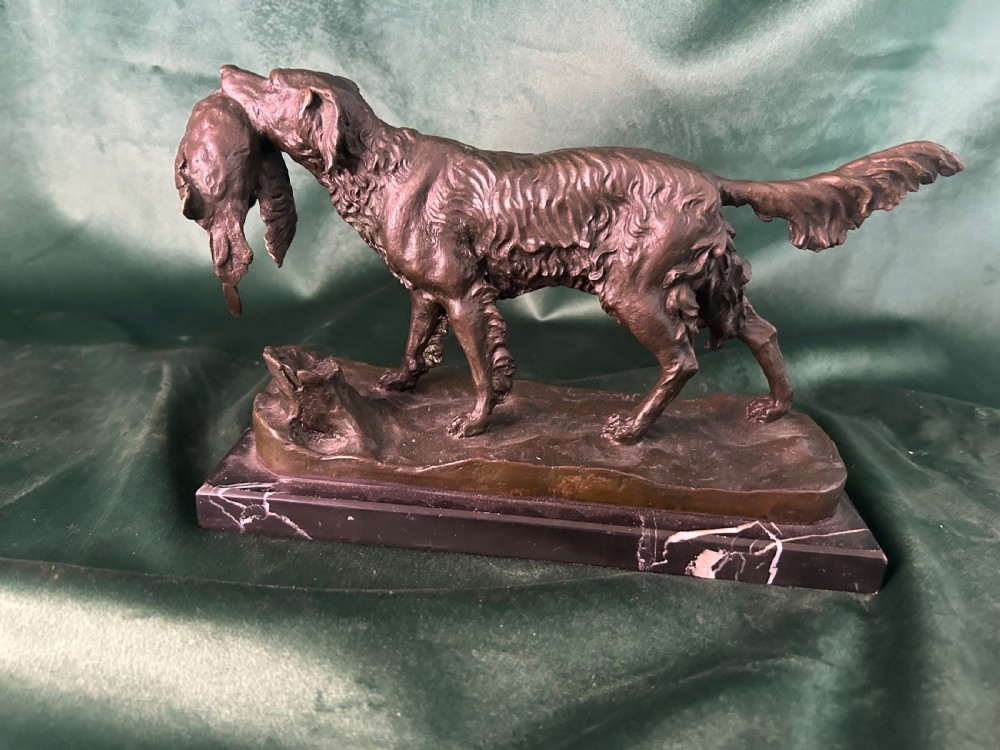 A cast bronze figure of a hunting dog holding a game bird in its jaws, set on a naturalistic ground base and mounted on a black veined marble plinth.