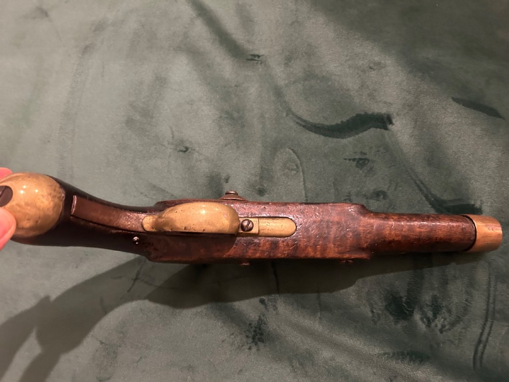 An antique single-barrel percussion pistol featuring a carved walnut stock, a metal hammer mechanism, and brass fittings along the trigger guard and butt cap. View VI