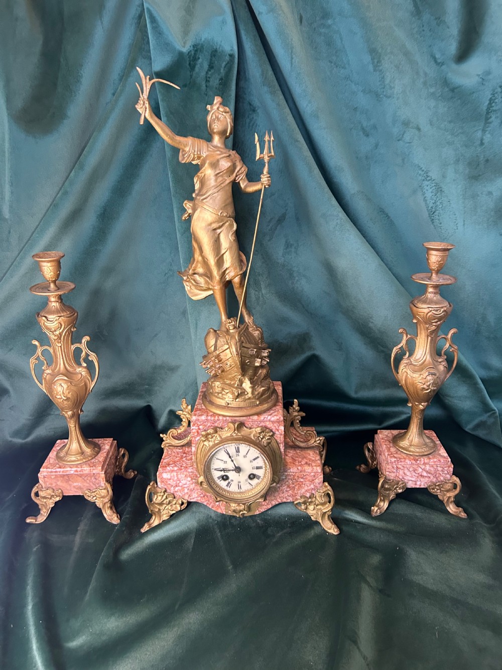 A three-piece French clock garniture set featuring a central mantel clock topped with a gilded classical figure on a pink marble base, flanked by two matching gilded side urns.