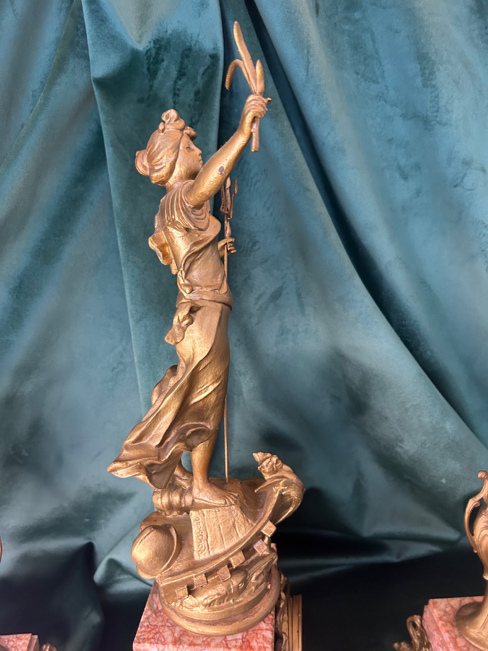 A three-piece French clock garniture set featuring a central mantel clock topped with a gilded classical figure on a pink marble base, flanked by two matching gilded side urns. View V