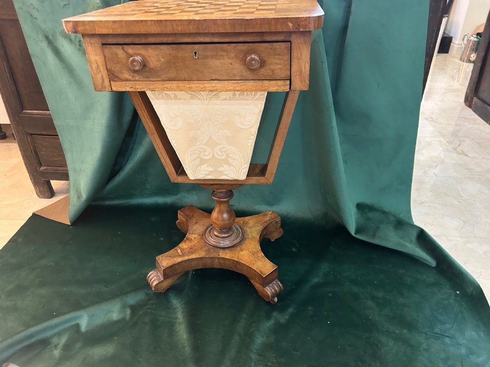 A Victorian warm brown wood games work table featuring an inlaid chessboard top, a single drawer, a damask fabric sewing well, and a turned pedestal base.